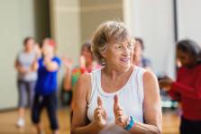 Pretty senior woman claps as she learns a new dance move during dance lessons at local senior center. The women is watching the instructor. People are dancing in the background.